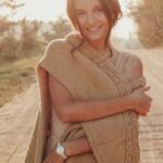 Smiling woman wrapped in a knitted sweater standing on a sunlit dirt road.